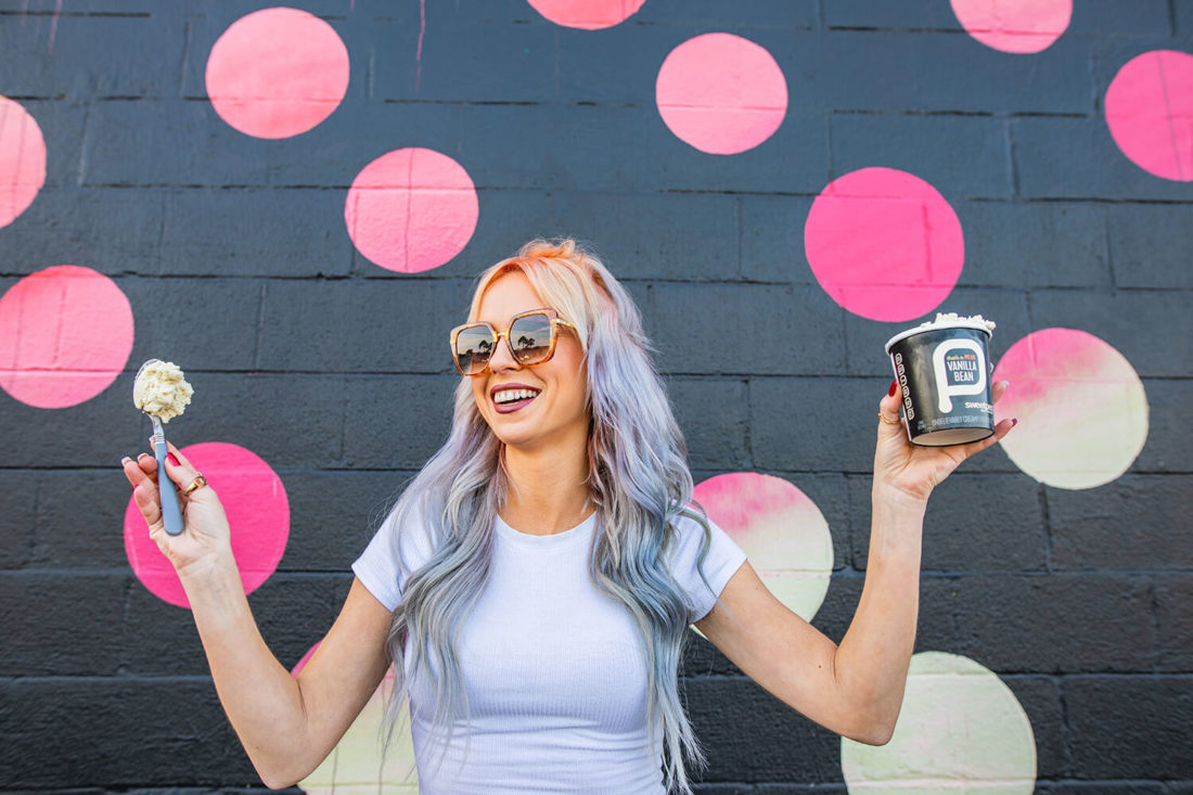 Edgy woman standing in front of a black and pink mural holding a pint of sweetpea plant-based ice cream.