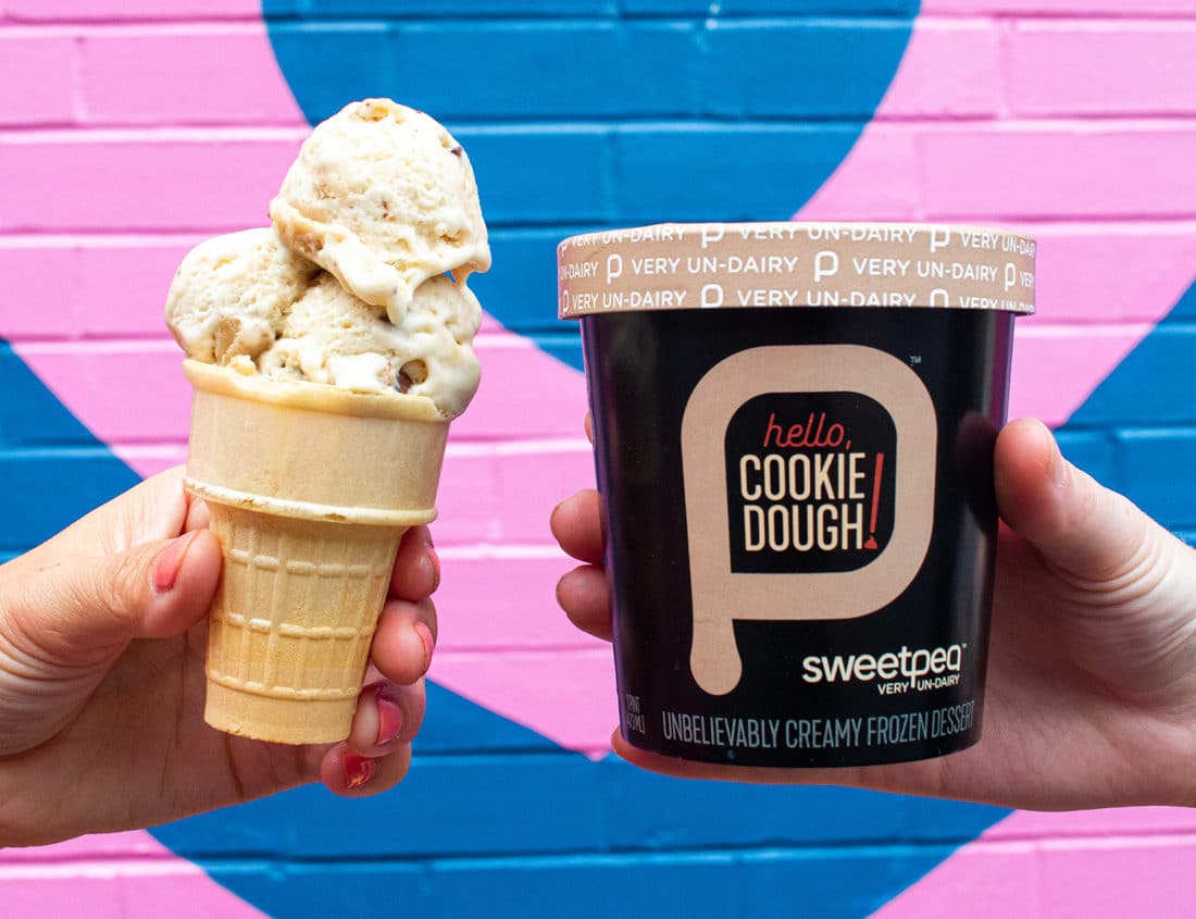 Pint of SweetPea Hello, Cookie Dough! Held next to an ice cream cone with the ice cream in it. Both are in front of a brick wall with a blue and pink heart mural on it.
