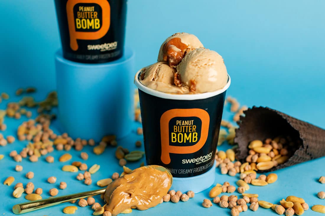 This is a photo os SweetPea plant-based ice cream flavor Peanut Butter Bomb. It shows an open pint against a blue background. Scattered all around are peanuts.