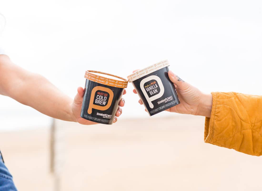 Two hands holding a pint each of SweetPea plant-based ice cream, clicking them together like drinking glasses.